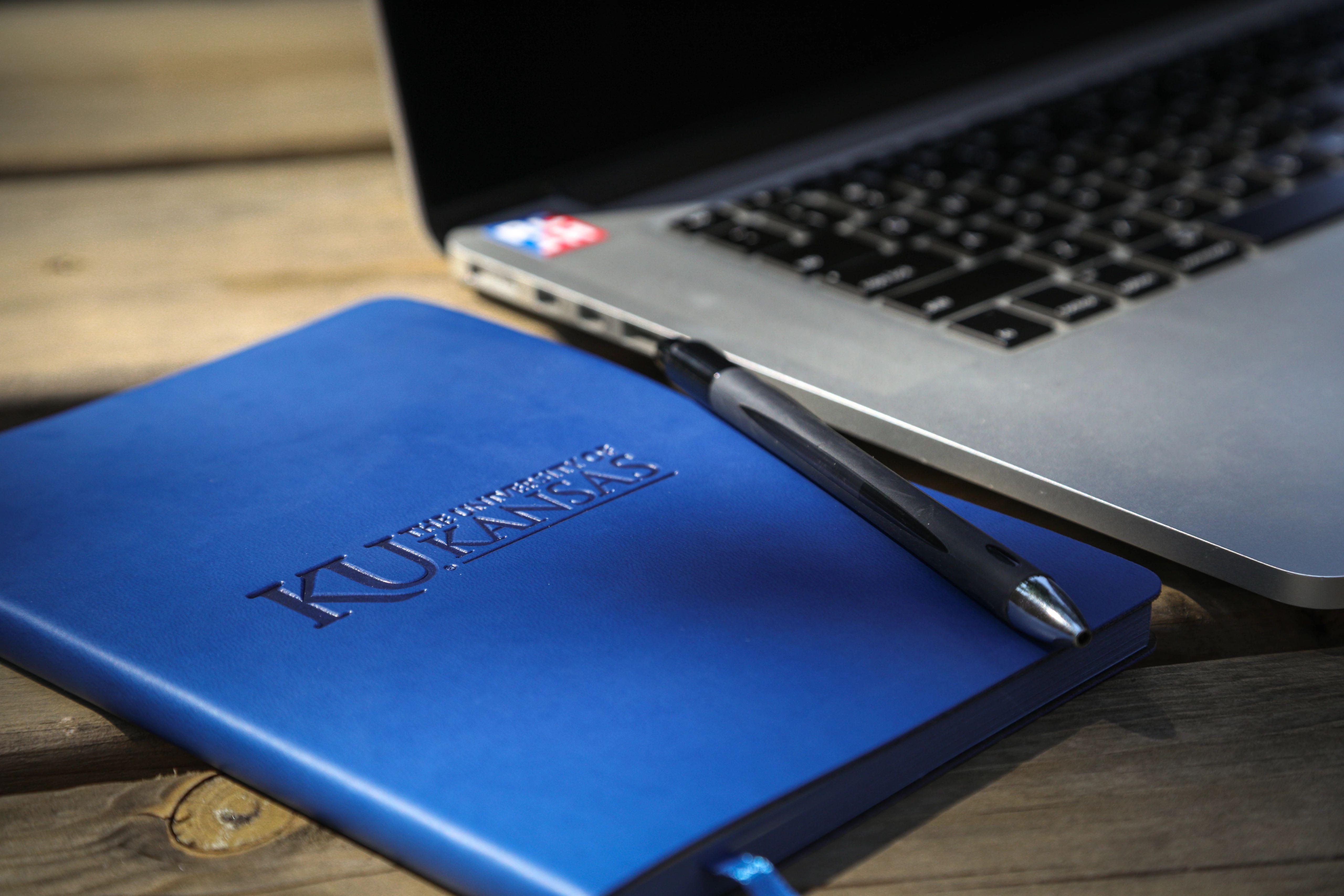 Blue KU notebook with pen on top next to laptop