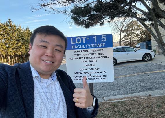 Professor Zhang giving a thumbs up standing in front of the Blake Hall parking lot.